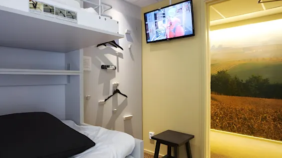 A modern, compact room featuring a bunk bed with visible instructions, a wall-mounted TV, and hangers. A doorway reveals a nature-themed wall.