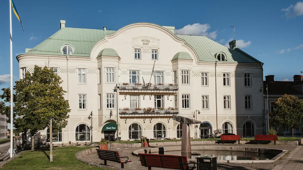 A grand white building with a green roof and balconies, labeled HOTELL POST and Hotel Post, stands behind a park with benches and a pond.