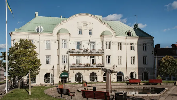 A grand white building with a green roof and balconies, labeled HOTELL POST and Hotel Post, stands behind a park with benches and a pond.