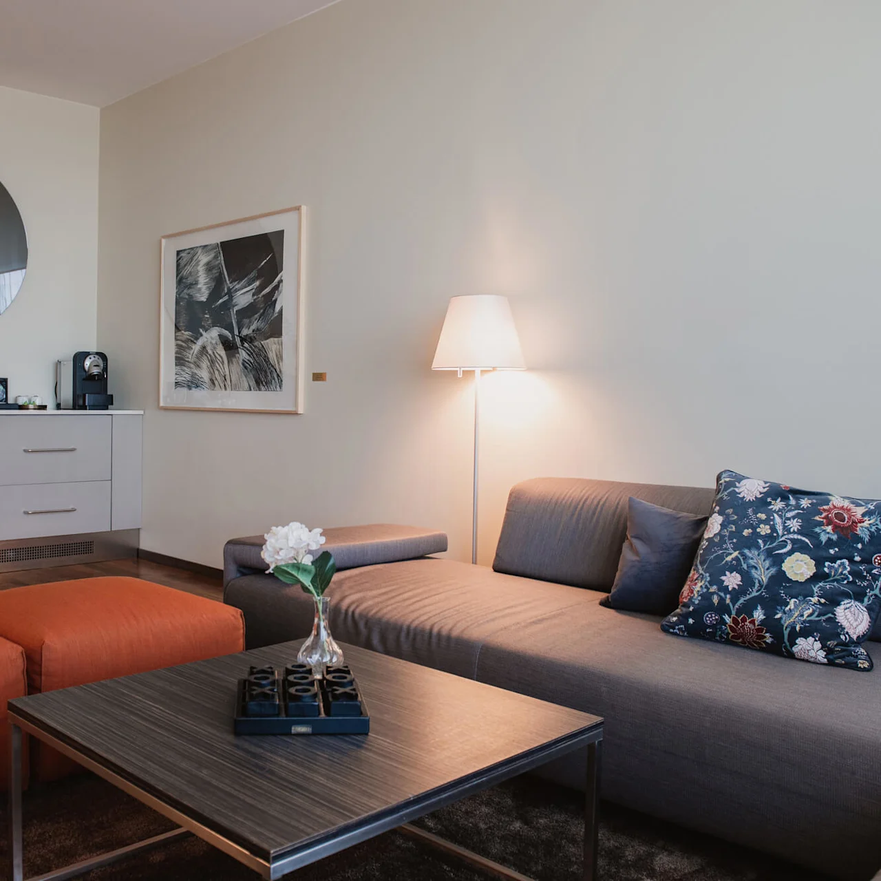 A gray sofa with floral cushions sits in a modern living room, accompanied by a table with a flower vase. Nearby, two orange poufs stand beside a cabinet with decor and a mirror.