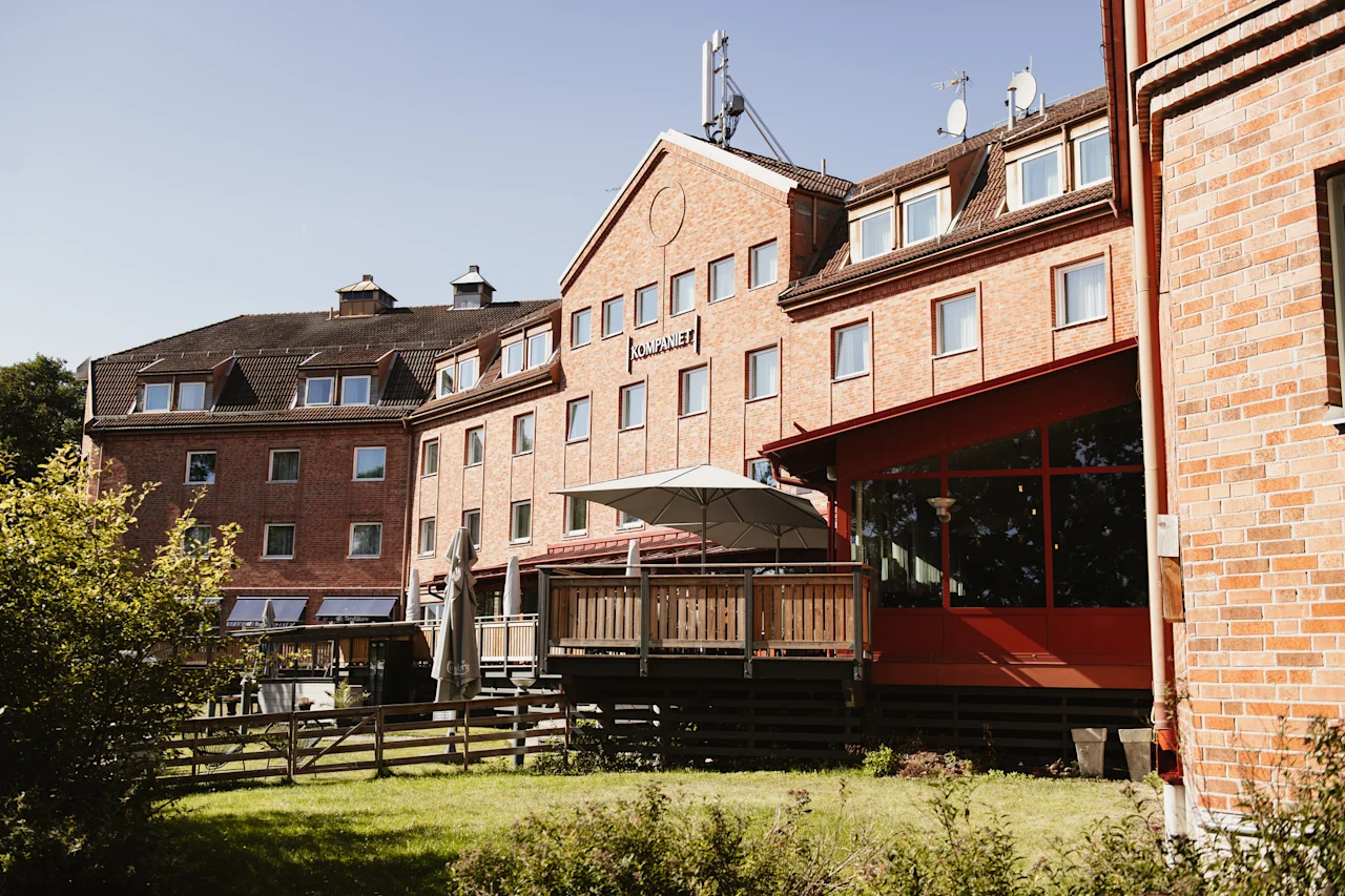 En tegelbyggnad med en skylt där det står KOMPANIET, med en utomhuspatio i trä med parasoller och en grön gräsmatta framför.