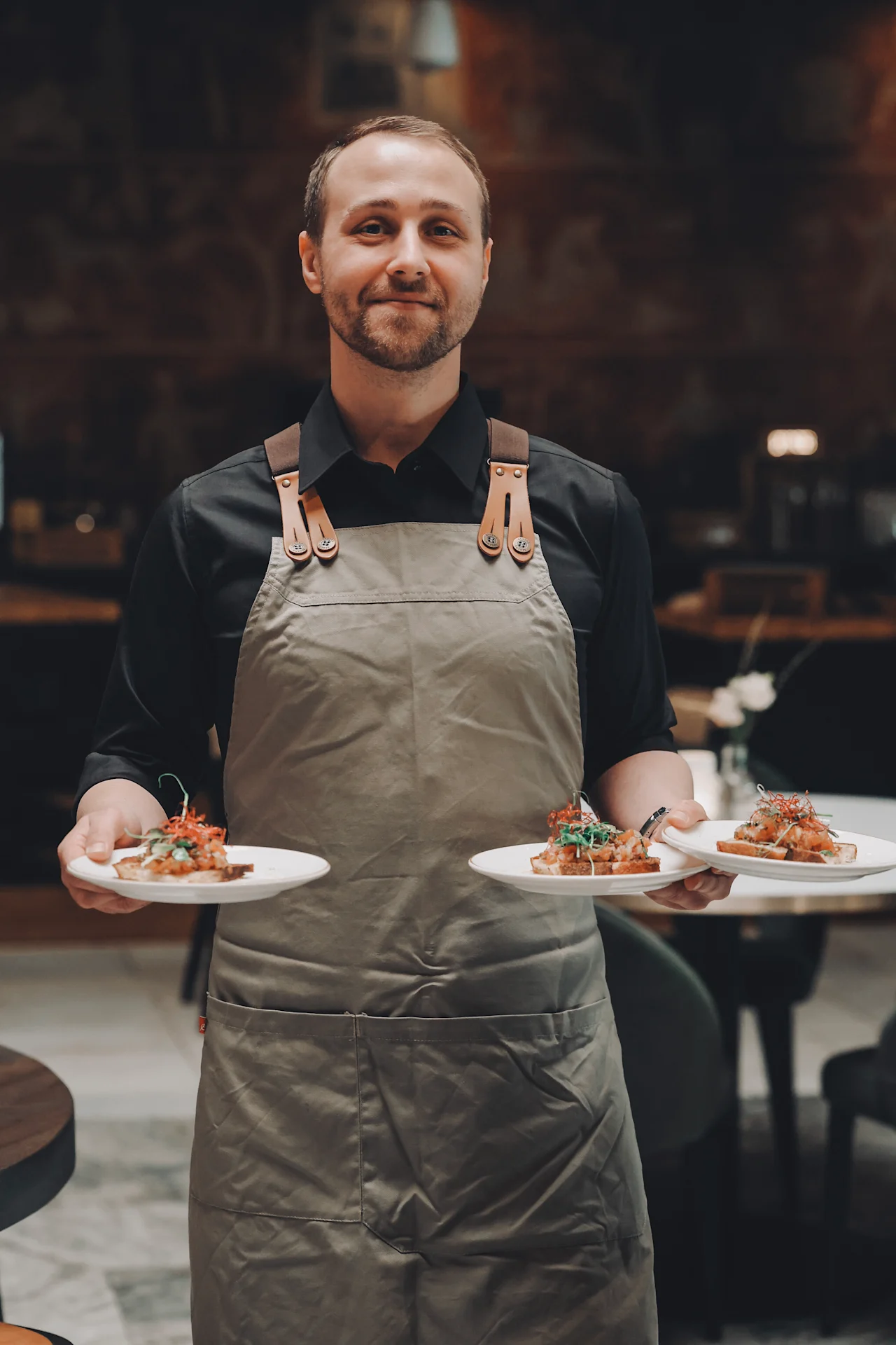 A waiter carries three plates of appetizing bruschetta, wearing a gray apron in an upscale restaurant setting with dark, elegant decor.