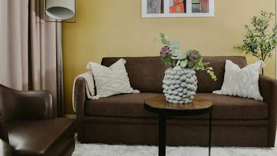 A brown sofa with white cushions sits against a yellow wall. A vase with flowers is on a wooden table. A modern floor lamp and beige curtains complete the cozy room.