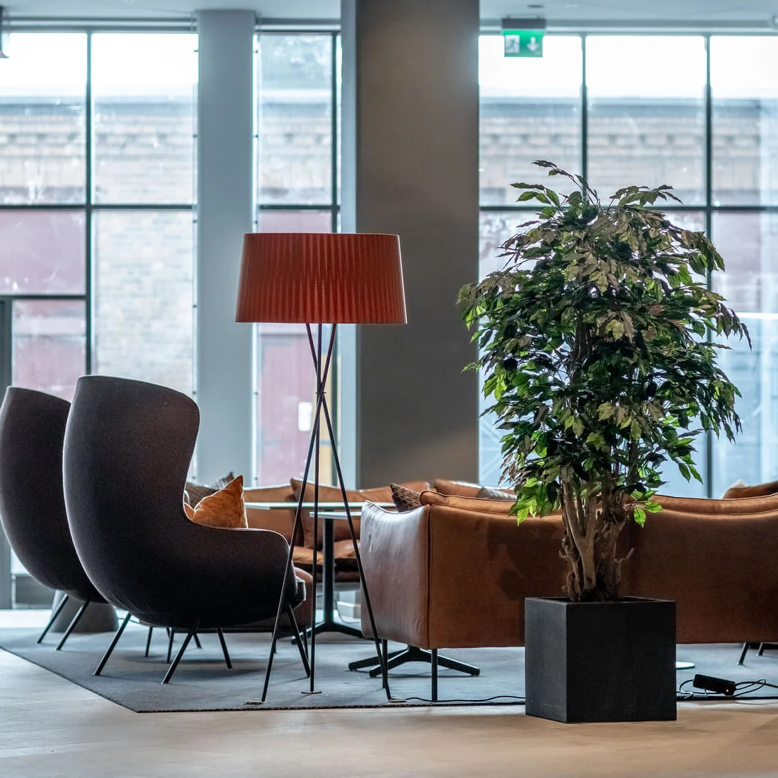 Et hyggeligt siddeområde med mørke stole omgiver et sofabord. En stor potteplante og en gulvlampe pynter rummet. Store vinduer afslører en murstensvæg.