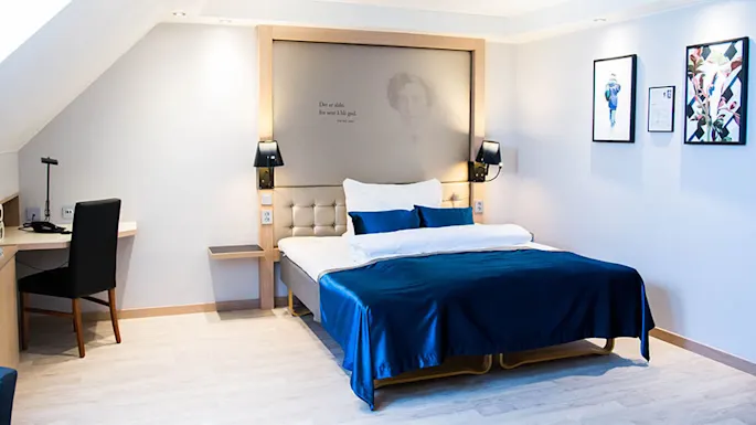 Bed centered beneath backlit headboard with blue bedding; wall on right has framed art. Text reads: “Det er aldri for sent å bli god.” Desk and chair on left.