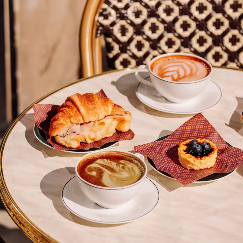 Kaffe og kager på et rundt cafébord i sollys, med en dekorativ stol i baggrunden.