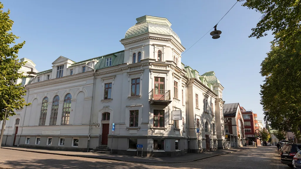 Upea rakennus, Home Hotel Victoria, jossa on erottuva katto ja arkkitehtonisia yksityiskohtia, seisoo mukulakivikadulla kirkkaan sinisen taivaan alla.