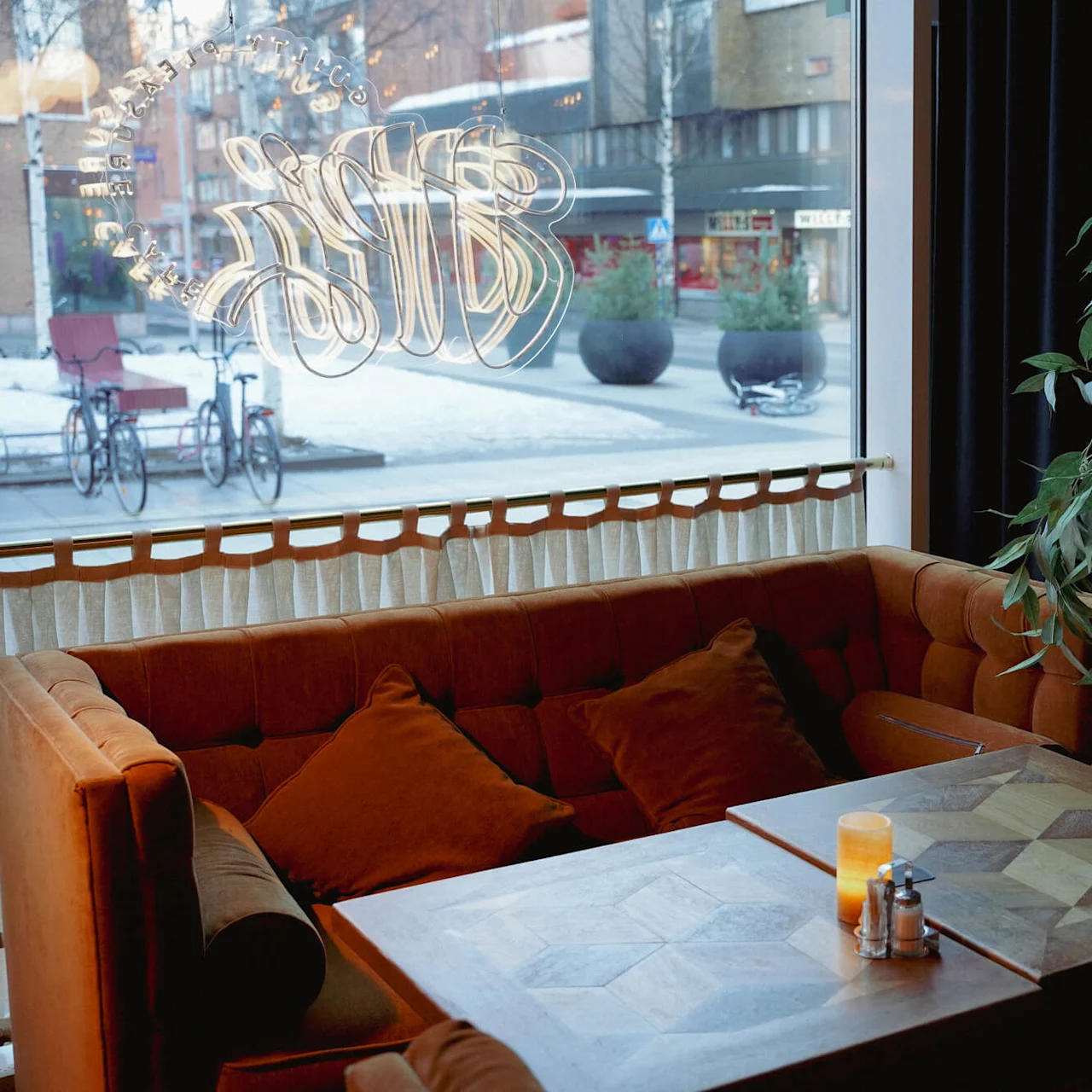 Et hyggeligt caféhjørne med en fløjlsofa og bord, set gennem et vindue med udsmykket skrift, der reflekterer bygaden udenfor.