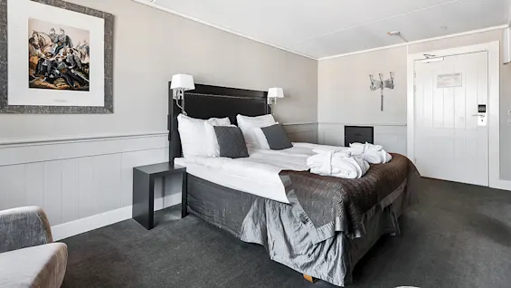 Neatly made bed with grey and white bedding in a tidy, modern bedroom setting with a wall-mounted artwork and dark carpet.
