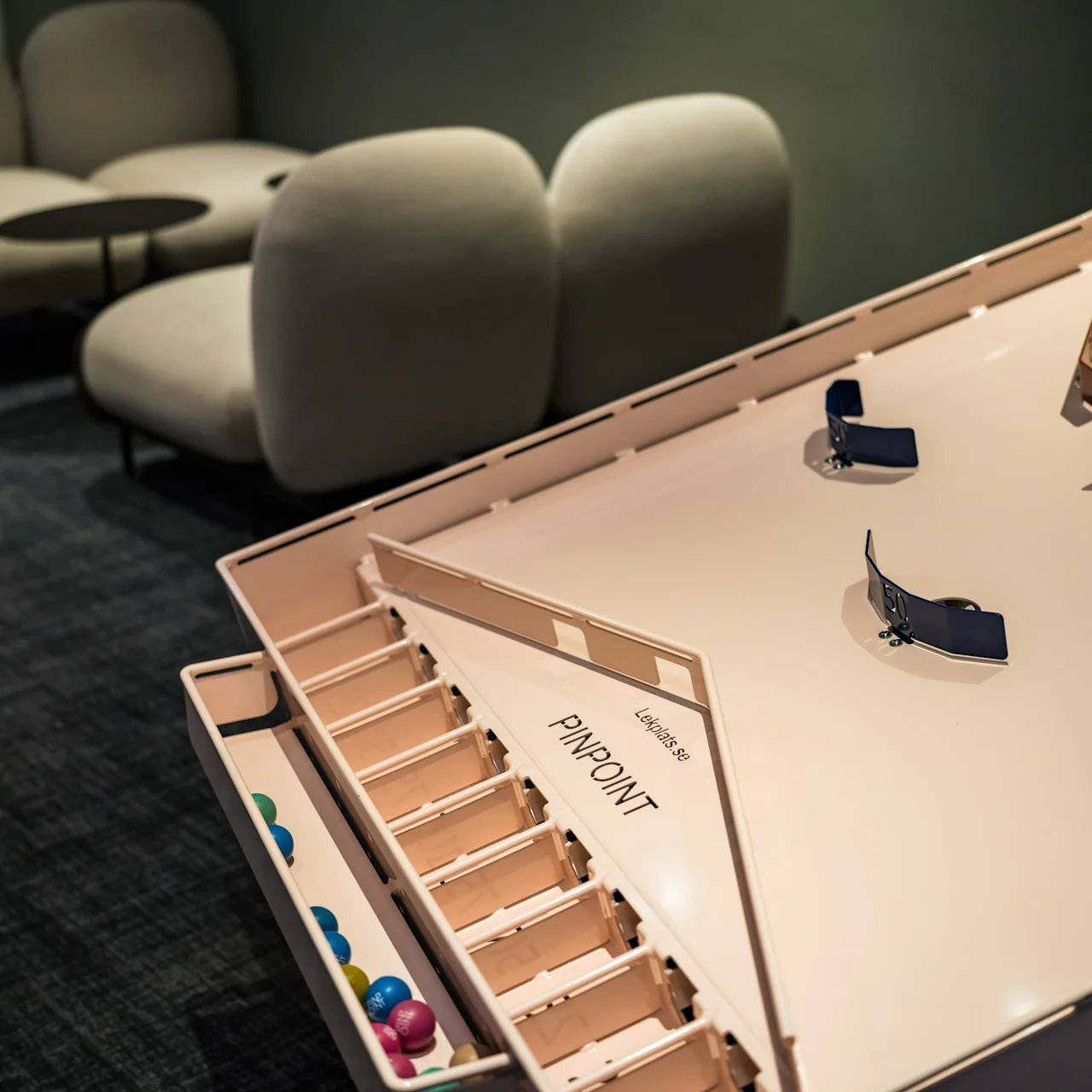 A tabletop game board labeled "Miniature PinPoint" in a dimly lit room, with colorful small balls and curved tracks. Background has plush seating and small tables.