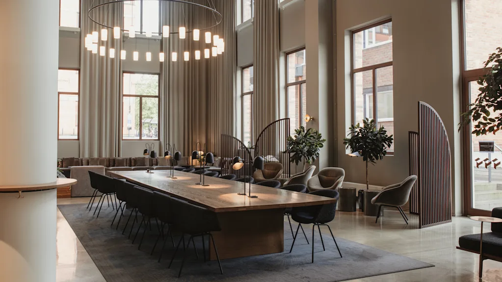 A modern and spacious hotel lobby with a long communal table, lounge seating, and large windows. Home Hotel Tapetfabriken.