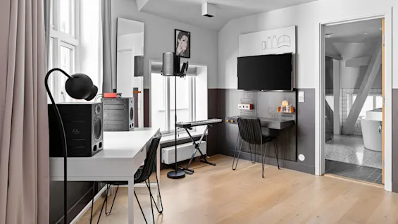 Et moderne arbeidsområde har et skrivebord med lampe og høyttaler til venstre, et tastatur i midten og en veggmontert TV overfor et bad med frittstående badekar.