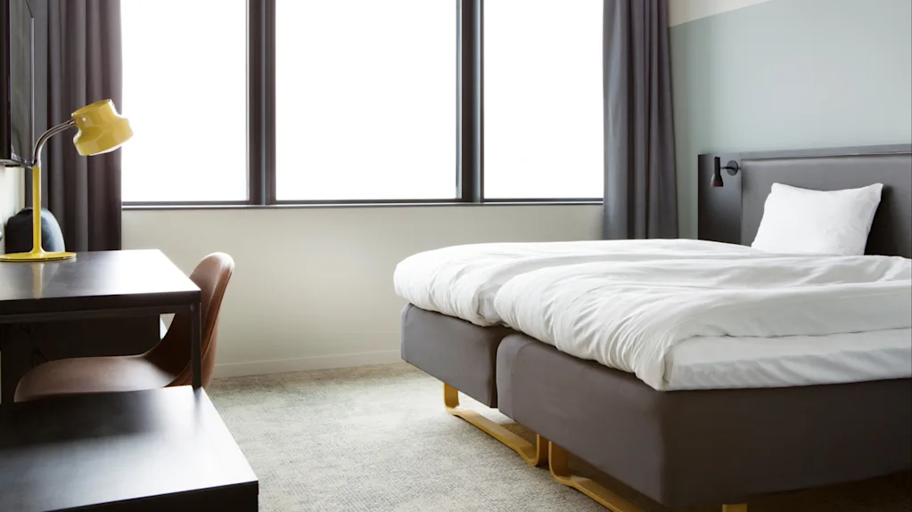 A neatly made bed with white linens rests near large windows in a simply furnished room featuring a desk with a yellow lamp.