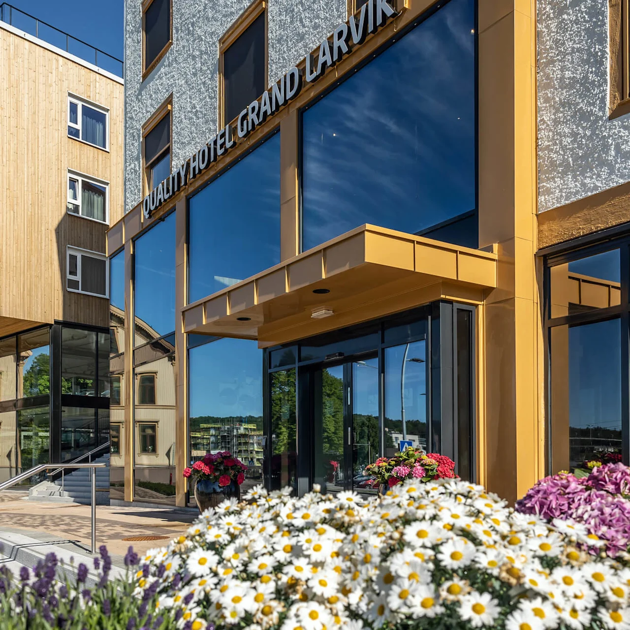 En bygning med moderne træ- og glasdesign, mærket "QUALITY HOTEL GRAND LARVIK," omgivet af livlige blomster og solskin, afspejler et roligt nabolag.