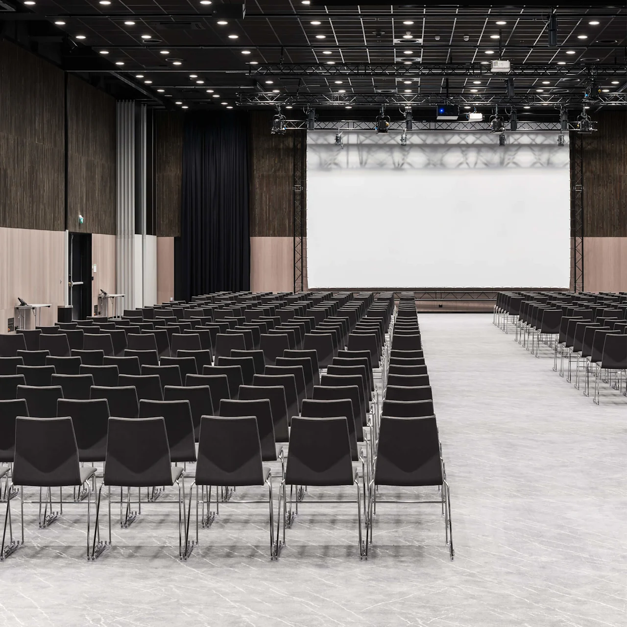Rader med svarte stoler vender mot en stor, tom projektorskjerm i et romslig, godt opplyst konferanserom med høyt tak med spotlights.