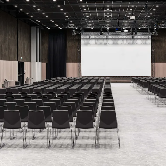 Rader med svarte stoler vender mot en stor, tom projektorskjerm i et romslig, godt opplyst konferanserom med høyt tak med spotlights.