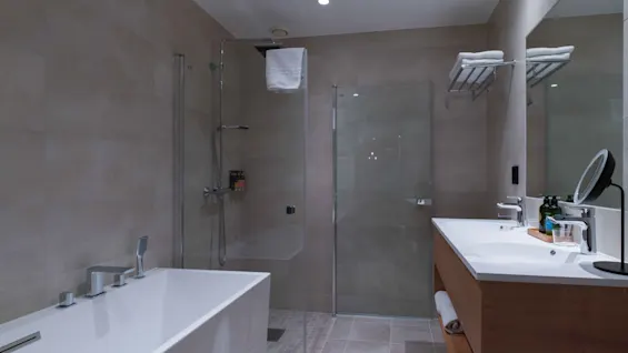 A modern bathroom features a white bathtub and a glass-enclosed shower. A wooden vanity with double sinks holds toiletries, and soft lighting enhances the clean, minimalistic design.