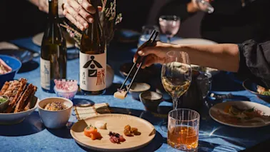 En hand använder ätpinnar för att ta sushi från en träplatta. En annan hand håller en sakeflaska med japansk text. Olika rätter och glas omger det blå bordet.