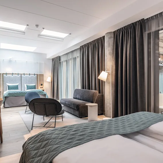 Et moderne rom med en dobbeltseng og teal-sengetøy, en sofa og et skrivebord, omgitt av store vinduer og gardiner, opplyst av taklamper.