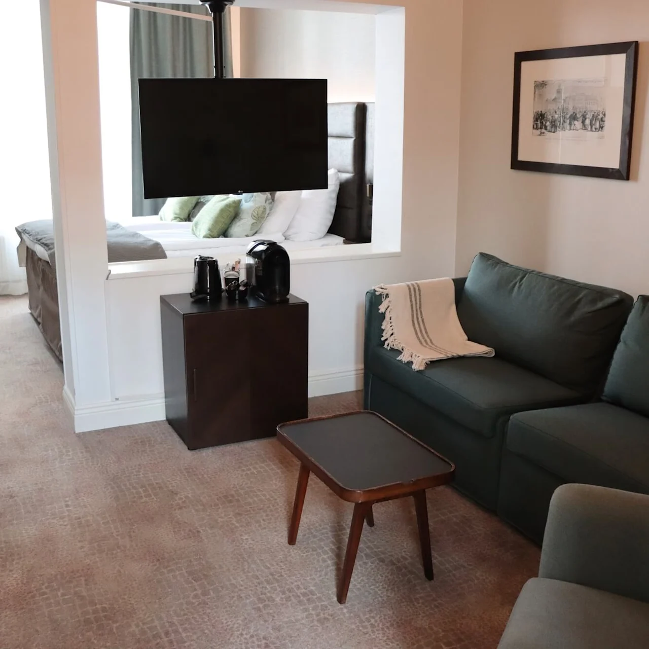 Ett hotellrum med en grön soffa, väggmonterad TV och en säng i det intilliggande rummet genom ett fönster i väggen.