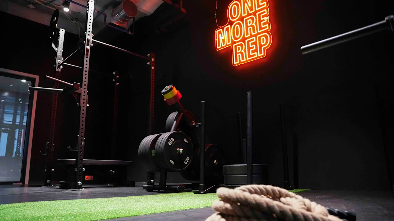 Et knebøystativ og treningsutstyr står ubrukt i et svakt opplyst treningsstudio. Neonskilt lyder "ONE MORE REP". Tykt tau ligger sammenrullet på grønt gress. Industrielt tak synlig.
