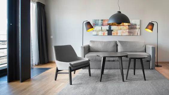 A gray sofa sits in a modern living room, paired with a gray chair and black tables. Floor lamps and a ceiling lamp illuminate the space, complemented by abstract wall art.