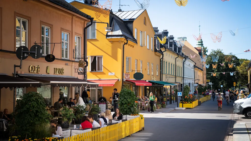 A vibrant street scene with people dining at outdoor cafes under butterfly decorations. Text reads CAFE LINNE.