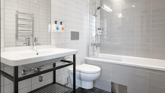 A modern bathroom with a wall-mounted sink below a mirror, next to a toilet and a bathtub with a showerhead. Dispensers labeled "SAPI" are mounted on tiles.