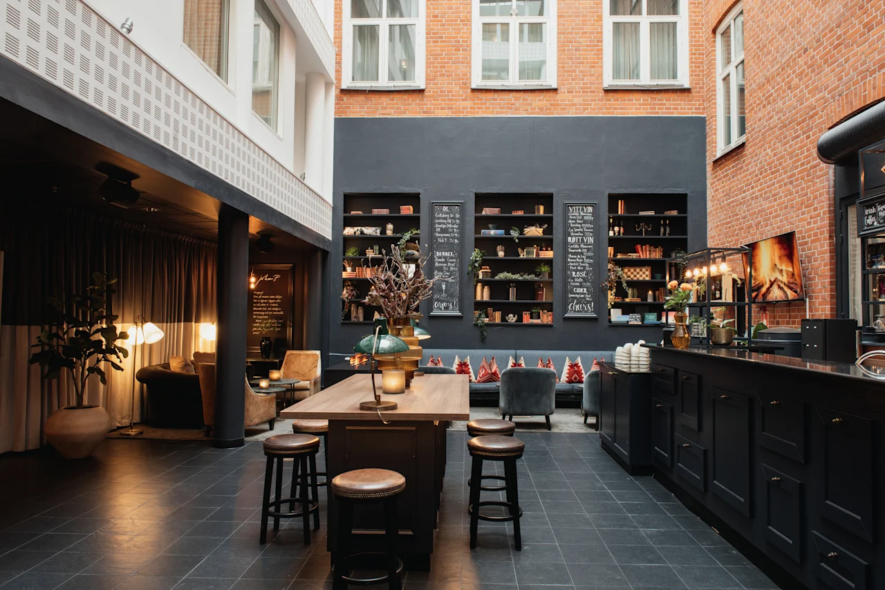 En varm og indbydende hotellounge med bar, fællesbord og siddepladser. Menupunkter som VITE VIN og ROSE er synlige.