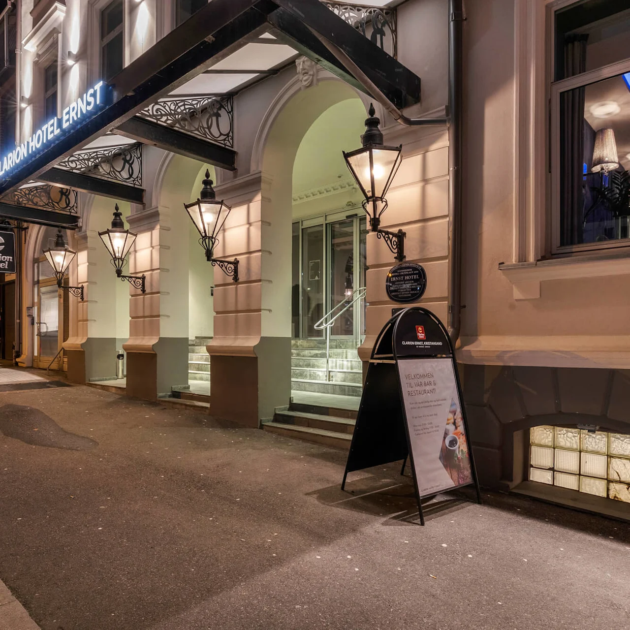 Clarion Hotels indgang belyst om natten, med arkitektoniske detaljer og skilte. I nærheden står en sandwichbræt med teksten: "Velkommen til vår bar & restaurant!" Gadeudsigt omfatter tilstødende bygninger.