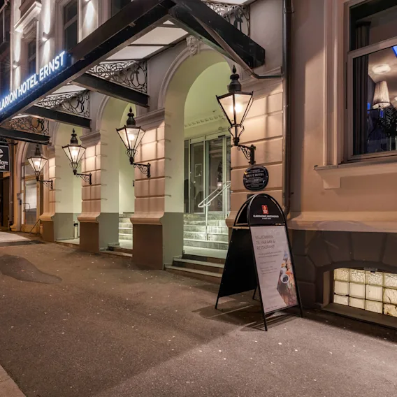 Clarion Hotel entrance illuminated at night, displaying architectural details and signage. Nearby, a sandwich board reads: "Velkommen til vår bar & restaurant!" Street view includes adjacent buildings.