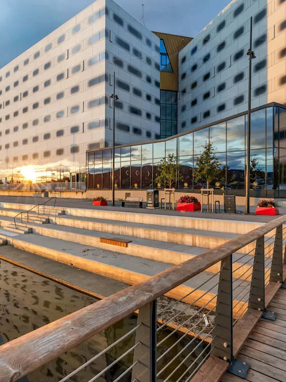 A modern building with reflective glass windows stands beside a stepped promenade by the water, under a cloudy sky. Red planters dot the area, creating a lively atmosphere.