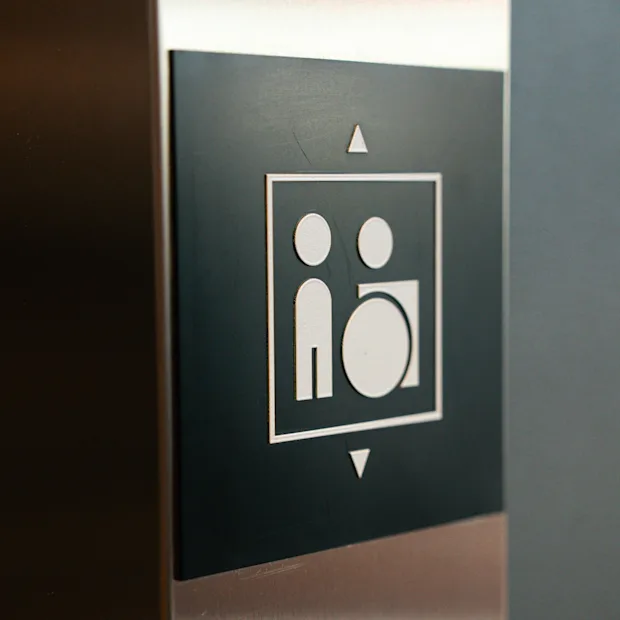 Hisskontrollpanel med en svart skylt som visar en vit ikon av två figurer, inramad av upp- och nedpilar.