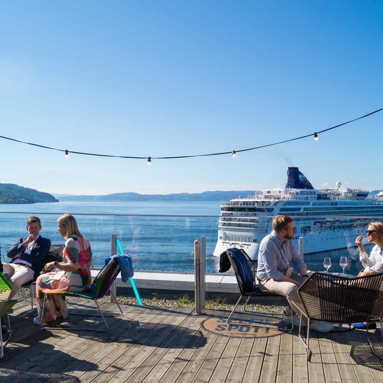 People sitting and talking at an outdoor cafe, overlooking a large cruise ship on calm blue water, with string lights above, surrounded by mountains under a clear sky.