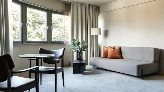 A gray sofa with orange pillows stands next to a tall lamp and a small table with flowers. Two black chairs are around a round table near large windows.