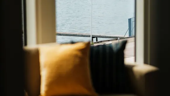 Et hyggeligt indendørs siddeområde med sofa og puder, der tilbyder en smuk udsigt over en sø med en ø og et flag.