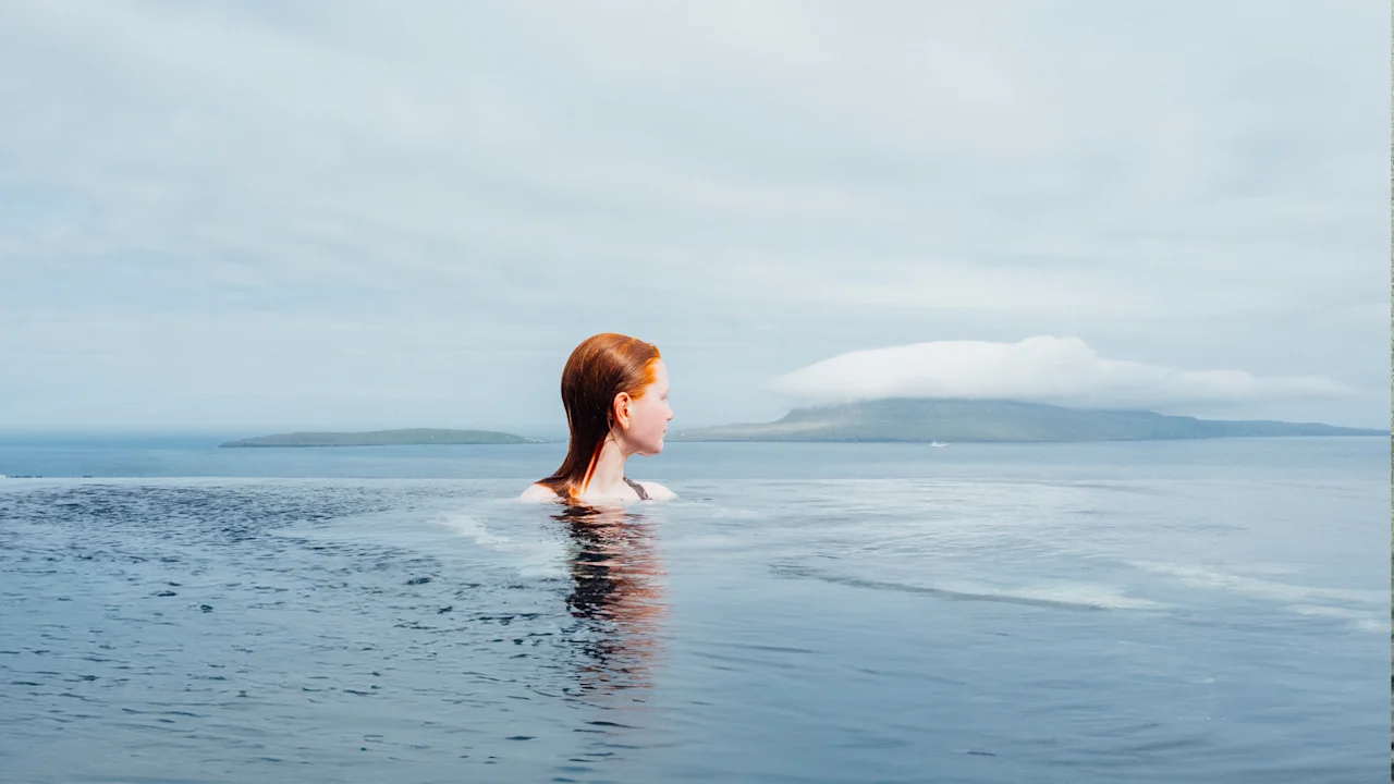 A person with red hair in an infinity pool, gazing out at a serene seascape with distant islands under a cloudy sky.