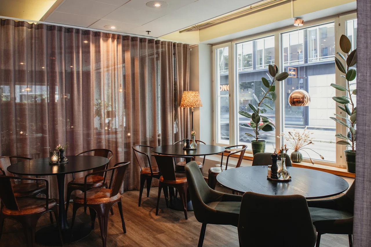 A cozy dining area at Home Hotel Uppsala with dark tables, comfortable chairs, and large windows showing ON and a city street.