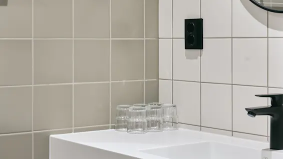 A modern bathroom with a white sink, black faucet, and round mirror. Glasses are on the counter, and toilet paper rolls are below.