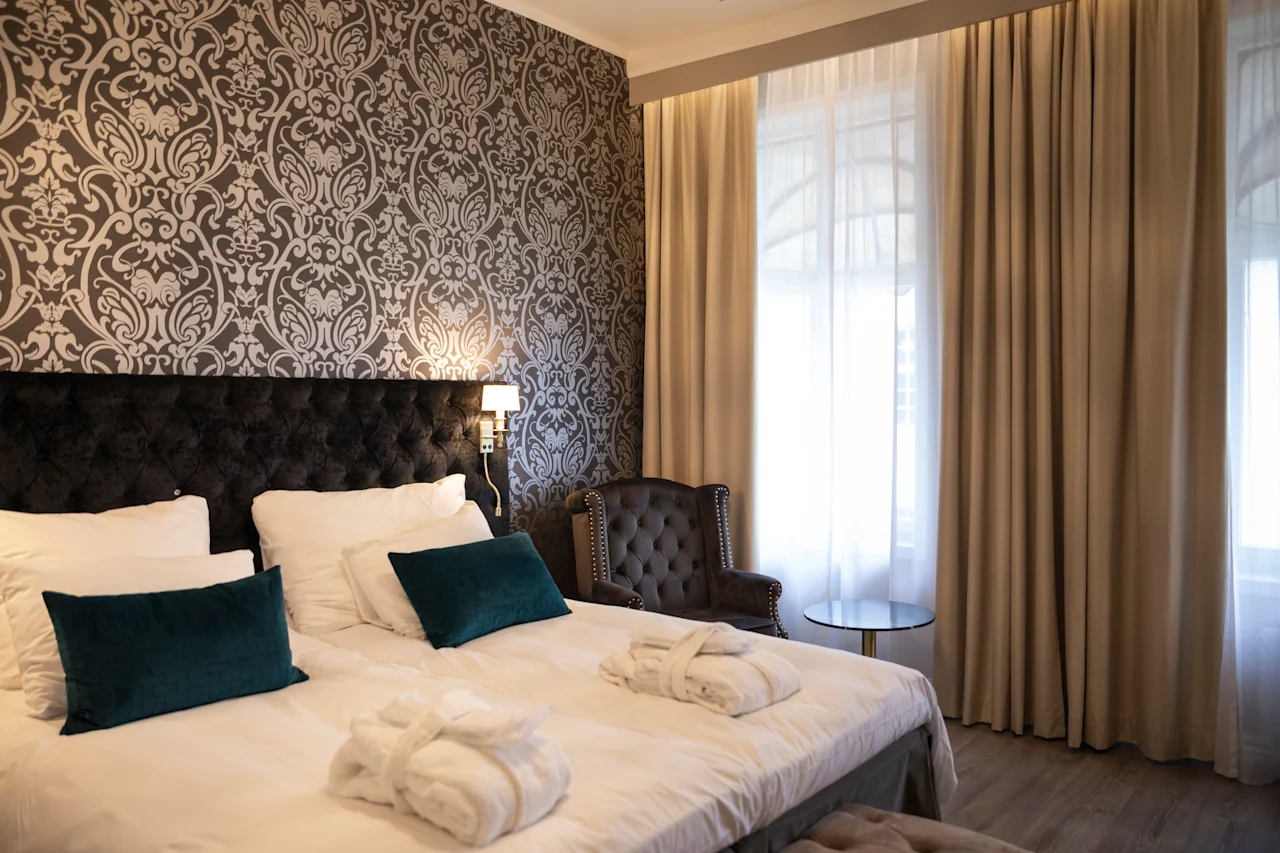 Et hyggeligt hotelværelse med en stor seng, elegant mønstret tapet og lyse vinduer med flagrende gardiner.