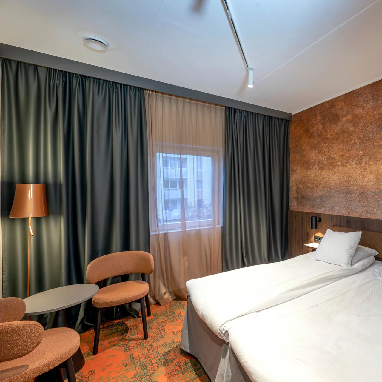 Double bed neatly made with white linens, positioned against a textured brown wall. Two chairs and a table sit by a window with dark curtains, creating a cozy atmosphere.