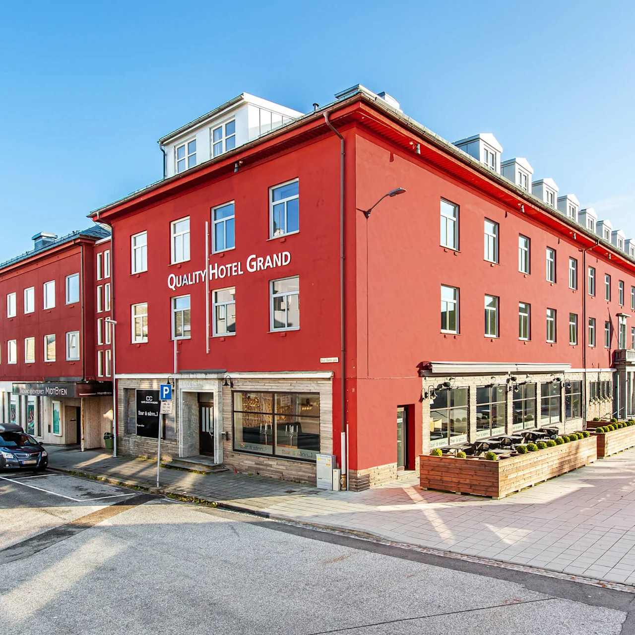 Quality Hotel Grand, en stor rød bygning, står på en gate med parkerte biler og klar himmel. Nær vann indikerer en kystnær setting.