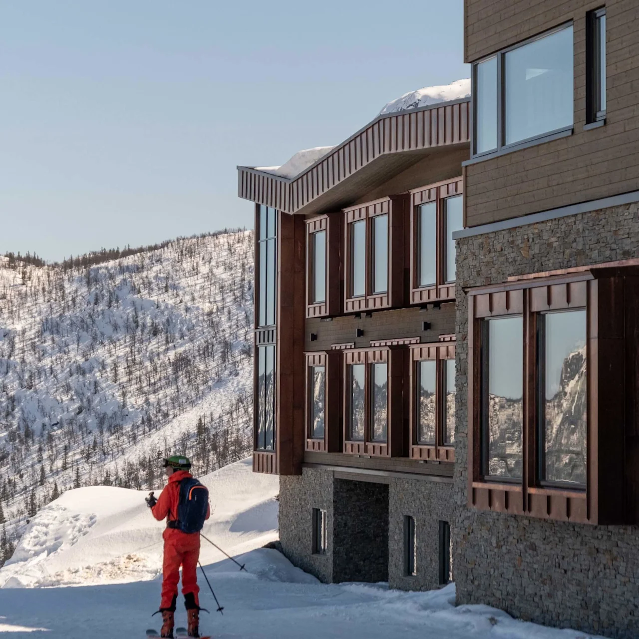 A skier in a red outfit stands near a modern building, overlooking a snowy mountainous landscape under a clear blue sky.