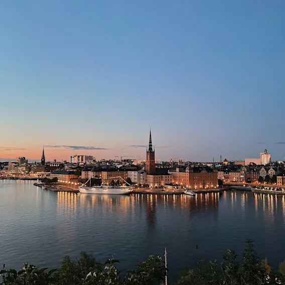 A city skyline at twilight, with illuminated buildings and spires reflecting on the water, under a soft, gradient sky.