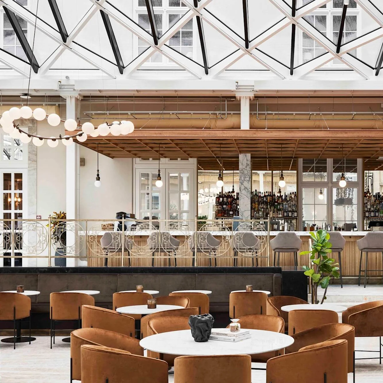 Bar with tall stools, under a modern glass ceiling, surrounded by brown lounge chairs and round tables, in a stylish, well-lit interior space.