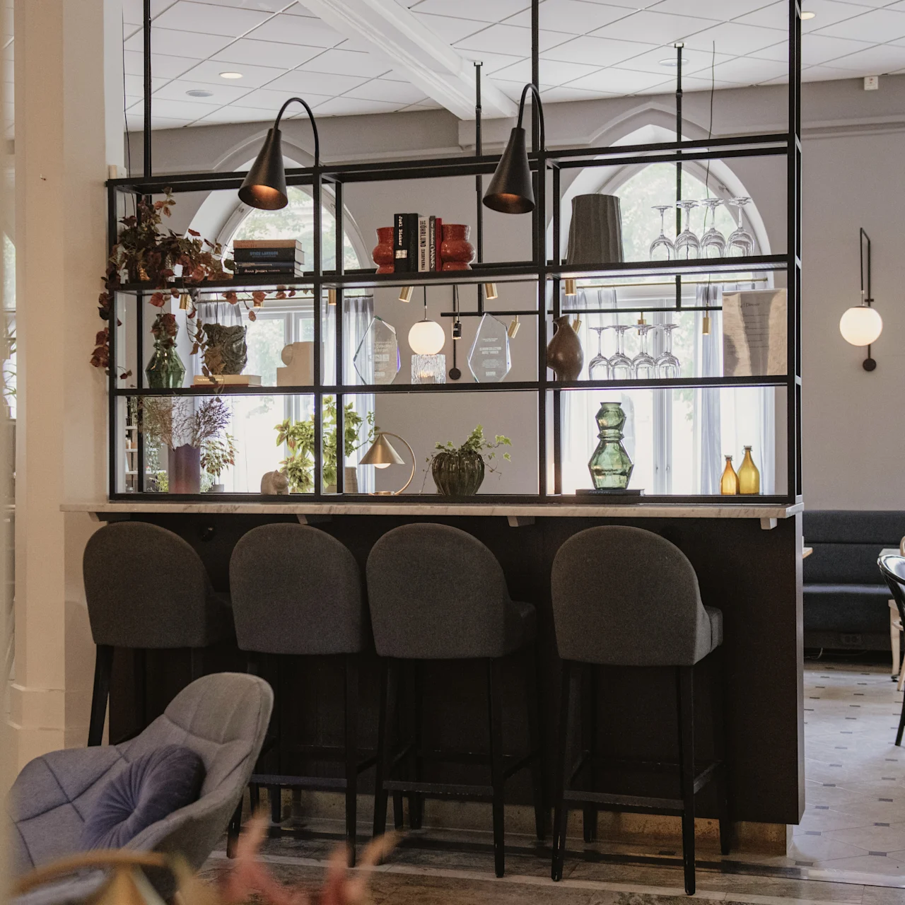 En bardisk med fyra högryggade stolar står mot en dekorativ hylla fylld med växter och vaser i ett stiligt kafé med modern belysning och välvda fönster.