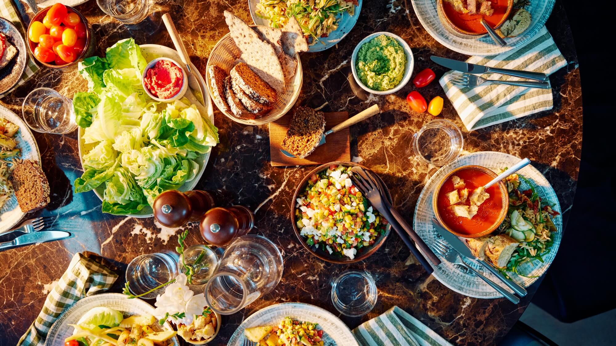 Bowls and plates filled with various salads, soups, and breads are arranged on a dark marble table, surrounded by cutlery, napkins, and glasses, creating a colorful dining setting.