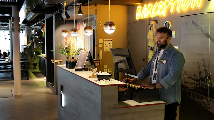 En smilende mand arbejder i en hotelreception med moderne dekoration og neonlys 'BARCEPTION'.