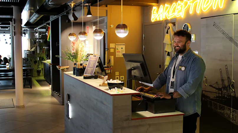 A smiling man works at a hotel reception with modern decor and neon 'BARCEPTION' sign.
