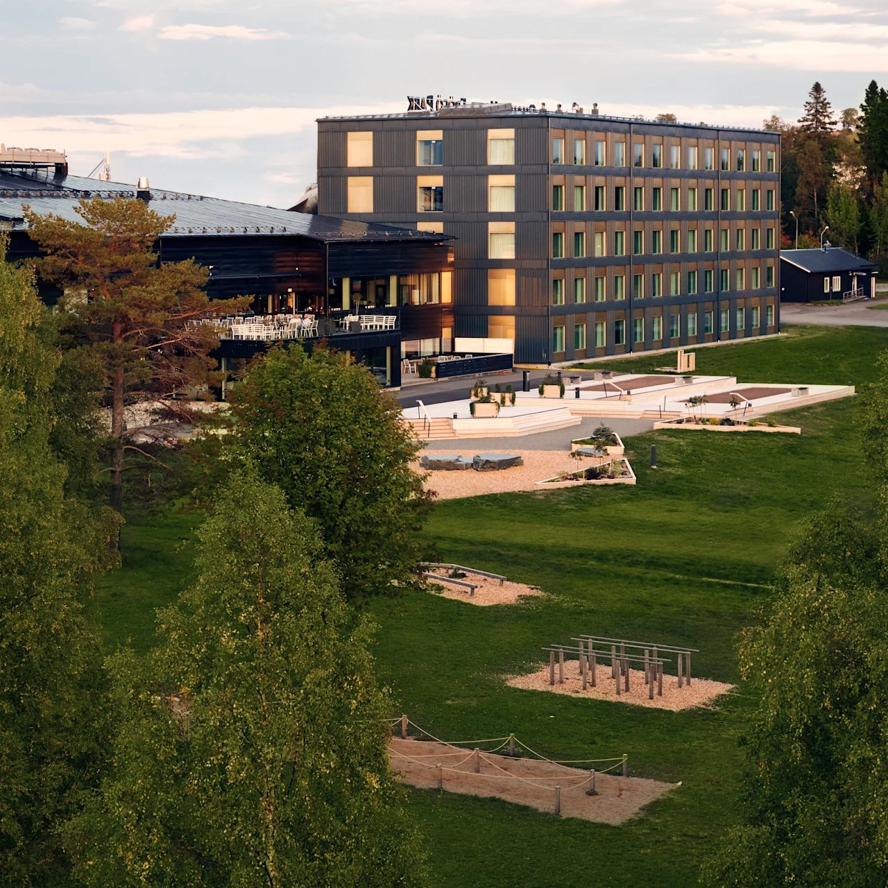 Ett modernt flerårigt byggnad står bredvid en grön park med olika utomhusträningsredskap, omgiven av stora träd. Himlen är delvis molnig, vilket tyder på en lugn sen eftermiddag.